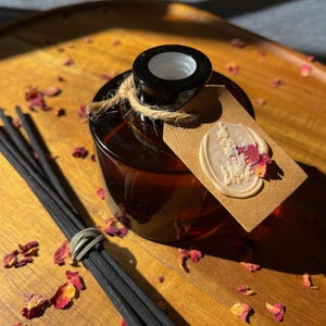 May include: A round amber glass diffuser bottle with a black neck and a bundle of black reed sticks. A brown paper tag is tied to the neck with twine, featuring a cream-coloured wax seal embossed with delicate floral patterns and pink petals.