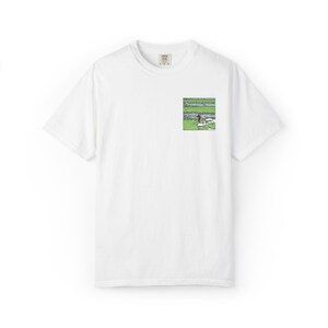 Może przedstawiać: Biały t-shirt z krótkim rękawem z małym, kwadratowym nadrukiem graficznym na lewej piersi. Grafika przedstawia stylizowany krajobraz z poziomymi pasami w kolorze zielonym i niebieskim, zawierający małą ciemną postać oraz białe kształty.