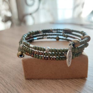 Puede incluir: Una pulsera de varias vueltas con cuentas de semillas en color verde oliva y bronce metálico. El diseño incluye pequeños espaciadores de metal plateado y un dije de pluma plateado que cuelga de un extremo de la estructura de alambre.