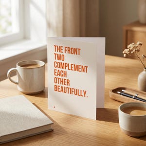 May include: A folded greeting card with a cream-coloured background and bold orange text that reads: THE FRONT TWO COMPLEMENT EACH OTHER BEAUTIFULLY. A small orange dot is printed at the bottom left corner of the card, which stands upright on a wooden surface.