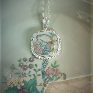 May include: A silver chain necklace with a square pendant featuring a hand-painted porcelain design of two birds in a floral setting.