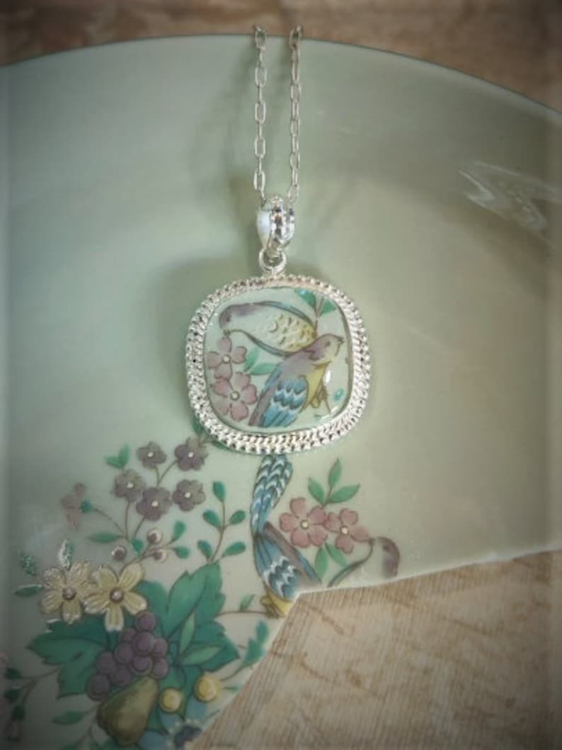 May include: A silver chain necklace with a square pendant featuring a hand-painted porcelain design of two birds in a floral setting.