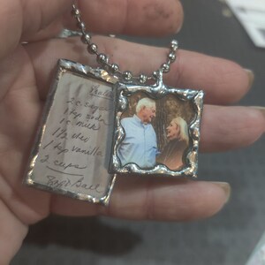 May include: A silver necklace with two charms. One charm is a small photo of a couple, and the other charm is a silver tag with handwritten text that reads "2 c. sugar, 1 1/4 c. soda, 1 c. milk, 1 1/2 oz. oil, 1 tsp vanilla, 3 cups, Soft Ball".