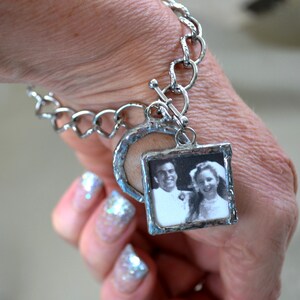 May include: A silver charm bracelet with a heart-shaped charm and a square charm with a black and white photo of a couple. The photo charm is attached to the bracelet with a silver chain.