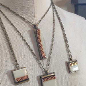 May include: Necklaces with cream-coloured pendants, each with a red and gold geometric design. The pendants are rectangular and square, with antique brass-coloured chains. The necklaces are displayed on a mannequin.