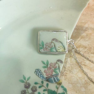 May include: A silver chain necklace with a square pendant featuring a hand-painted porcelain design of a bird perched on a branch with pink flowers. The pendant is set in a silver frame.