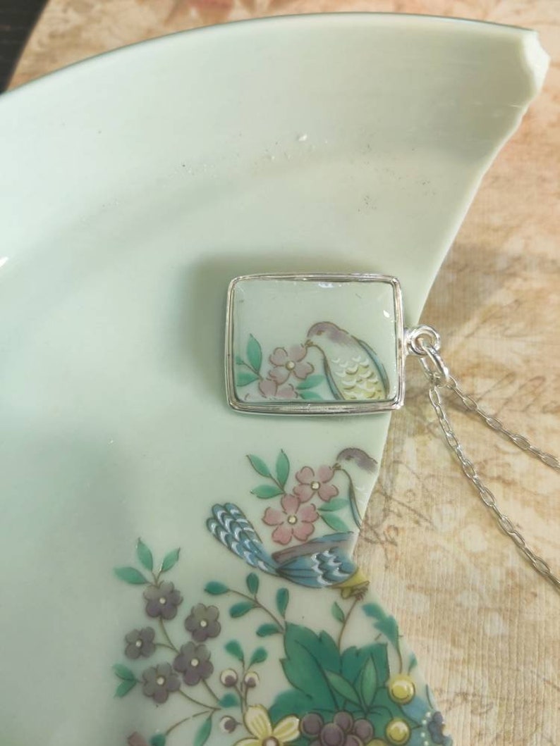 May include: A silver chain necklace with a square pendant featuring a hand-painted porcelain design of a bird perched on a branch with pink flowers. The pendant is set in a silver frame.