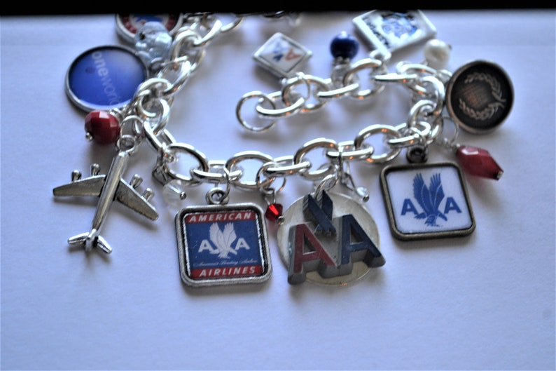 Puede incluir: Un brazalete de plata con varios abalorios, incluyendo un avi&oacute;n peque&ntilde;o, un abalorio cuadrado con el logotipo de American Airlines y un abalorio redondo con las letras "AA" en rojo, blanco y azul. El brazalete tambi&eacute;n tiene varias cuentas rojas y azules.