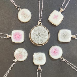 May include: A collection of seven silver-toned necklaces featuring white ceramic pendants. The center pendant is large and round with a rope-style border and grey swirl patterns, surrounded by smaller round and square pendants with floral and geometric designs.