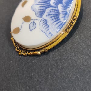 May include: Oval pendant necklace with a gold-tone chain. The pendant features a white porcelain design with blue floral patterns and gold accents, set in a gold-tone frame. The necklace is a classic jewelry piece.