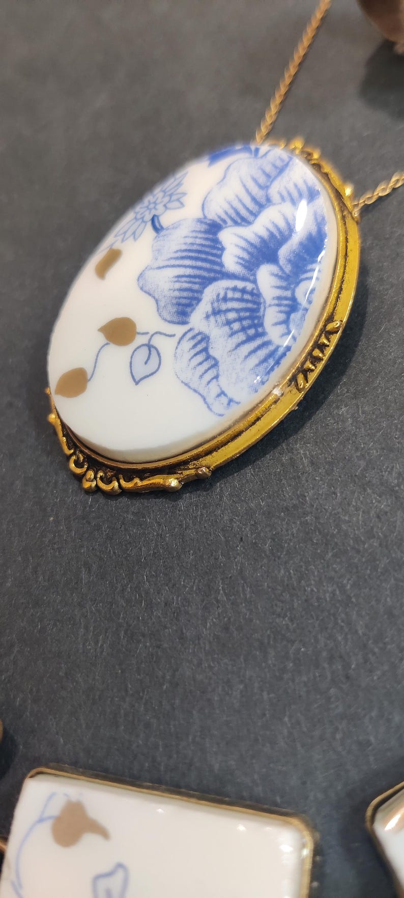 May include: Oval pendant necklace with a gold-tone chain. The pendant features a white porcelain design with blue floral patterns and gold accents, set in a gold-tone frame. The necklace is a classic jewelry piece.