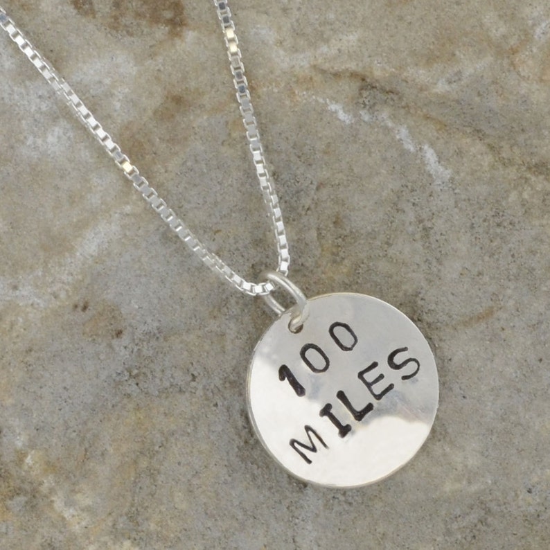 May include: Silver necklace with a round pendant that says "100 MILES" in black lettering.