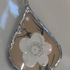 May include: A teardrop-shaped pendant made from a piece of tan ceramic with a white floral design and black branch accents. The ceramic fragment is encased in a silver-coloured metal solder frame and hangs from a delicate silver-coloured metal chain.