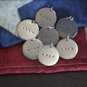 May include: Seven silver metal charms with the word "vote" engraved on each charm. The charms are arranged on a red, white, and blue American flag.