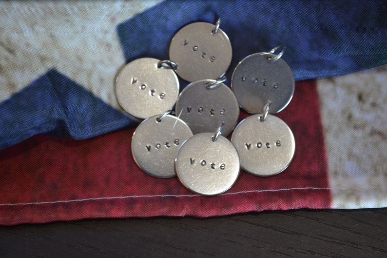 May include: Seven silver metal charms with the word "vote" engraved on each charm. The charms are arranged on a red, white, and blue American flag.