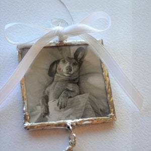 May include: A silver-framed ornament with a black and white photo of a dachshund dog inside. The ornament has a white ribbon and a silver bone charm hanging from it.