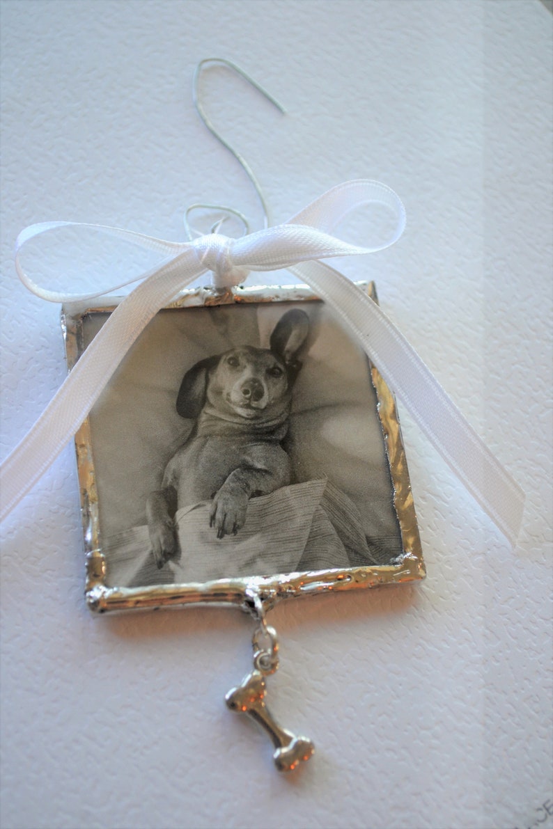 May include: A silver-framed ornament with a black and white photo of a dachshund dog inside. The ornament has a white ribbon and a silver bone charm hanging from it.