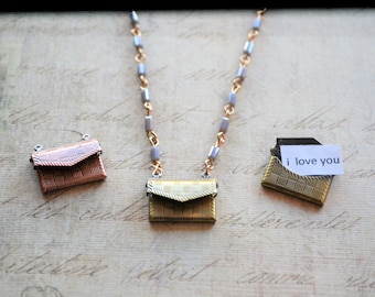 May include: Three gold-toned envelope-shaped pendants on a silver chain necklace. The pendant in the center is on the chain, while the other two pendants are on the surface below. The pendant on the right has a small white paper note inside that says "i love you".