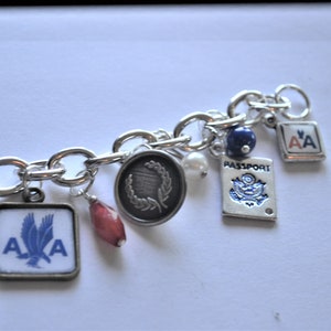 Puede incluir: Un brazalete de plata con varios dijes, incluyendo un dije de pasaporte, una cuenta roja, una perla, una cuenta azul y dos dijes con las letras "AA" en ellos.