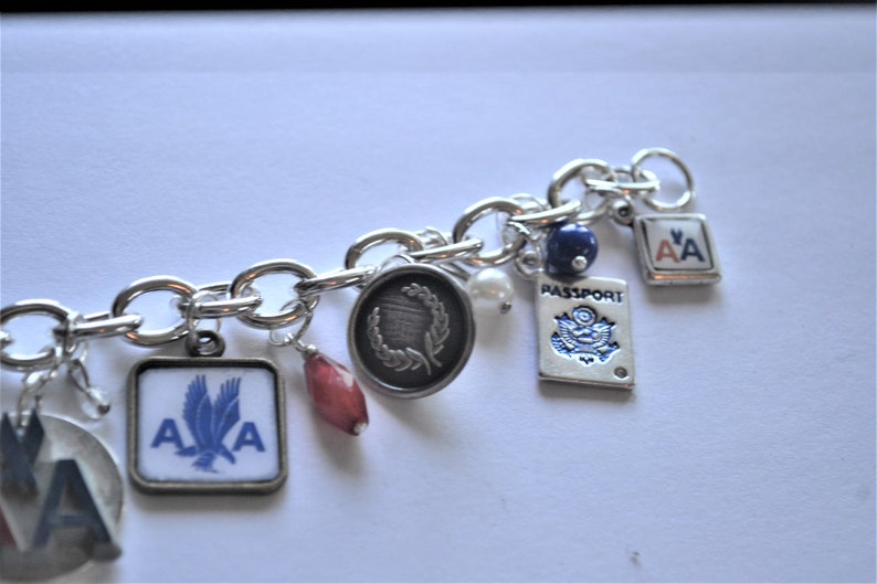 Puede incluir: Un brazalete de plata con varios dijes, incluyendo un dije de pasaporte, una cuenta roja, una perla, una cuenta azul y dos dijes con las letras "AA" en ellos.