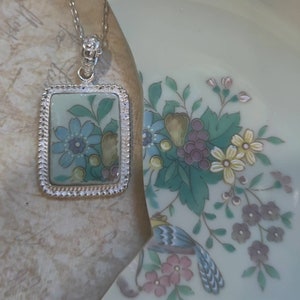 May include: A silver necklace with a rectangular pendant featuring a blue and green floral design. The pendant has a silver border with a decorative edge.