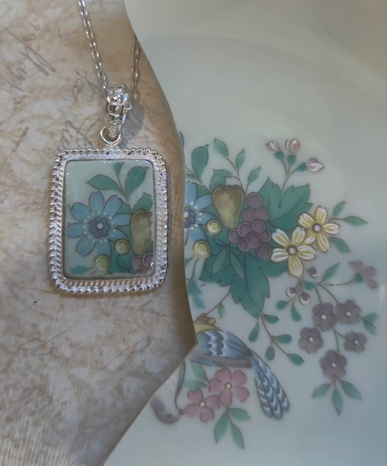 May include: A silver necklace with a rectangular pendant featuring a blue and green floral design. The pendant has a silver border with a decorative edge.