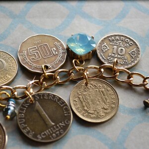 May include: A gold chain bracelet featuring a selection of vintage coins and a light blue gemstone charm. The coins include a 10 centimes coin from France, a 50c coin from Austria, a 10 francs coin from Morocco, and a 1 schilling coin from Austria.