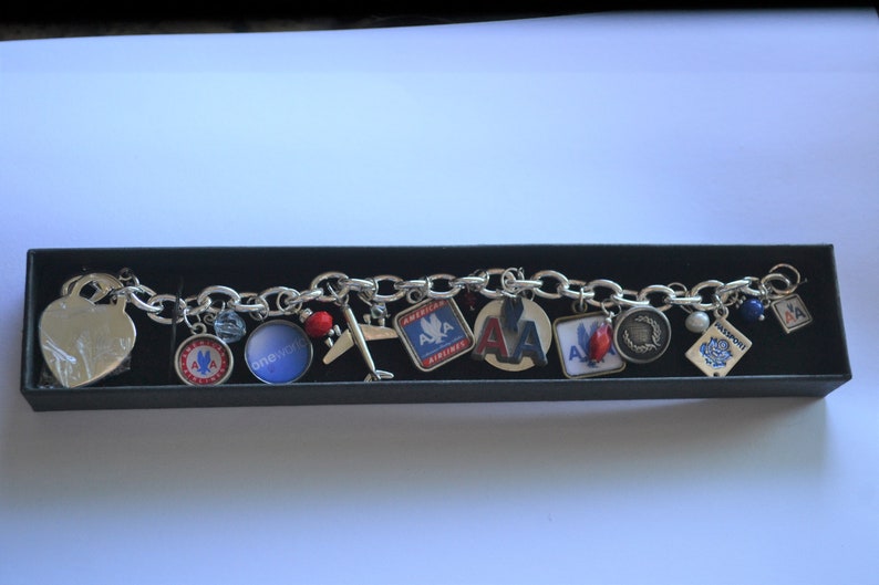 Puede incluir: Un brazalete de plata con varios dijes, incluyendo un coraz&oacute;n, un avi&oacute;n, un pasaporte y varios dijes con el logotipo de American Airlines. El brazalete est&aacute; en una caja negra.