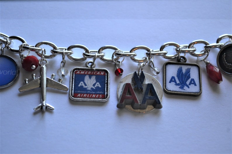 Puede incluir: Un brazalete de plata con varios dijes, incluyendo un avi&oacute;n peque&ntilde;o, un dije con el logotipo de American Airlines en rojo y azul, y un dije con el logotipo de American Airlines en rojo y azul con un &aacute;guila plateada.