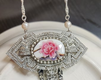 May include: A silver necklace with a floral pendant. The pendant features a pink flower on a white background, surrounded by a silver frame. The necklace has a silver chain with two pearl beads.