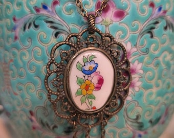 May include: A bronze necklace with a floral design pendant. The pendant is oval shaped and features a white porcelain cameo with a colorful floral design. The necklace has a chain with three dangling beads.