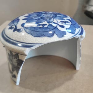May include: A broken blue and white porcelain lid with a floral design. The lid is cracked and chipped, but the floral design is still visible.