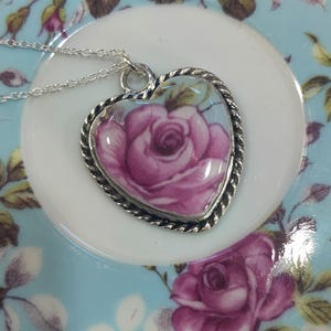 May include: A silver heart-shaped pendant necklace with a pink rose design. The pendant is on a blue and white floral background.