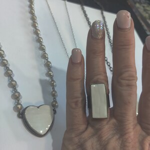 May include: A silver necklace with a white heart pendant and a silver chain. A white rectangular ring with a silver band is on a finger.