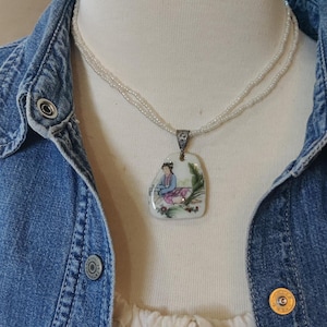 May include: A white porcelain pendant necklace with a delicate white bead chain. The pendant features a hand-painted image of a woman in traditional clothing sitting in a garden setting.