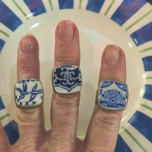 May include: Three square-shaped rings with blue and white porcelain designs. The rings are worn on the index, middle, and ring fingers of a hand.