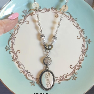 May include: A white pearl and crystal necklace with a vintage button charm and a cameo pendant. The pendant features a floral design on a white background. The necklace is displayed on a blue and white plate with a gold floral pattern.