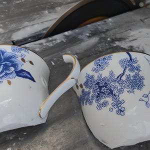 May include: Two broken white porcelain teacups with blue floral designs and gold trim. One cup features a large blue flower, while the other has a blue floral branch pattern. A circular saw blade is visible in the background.