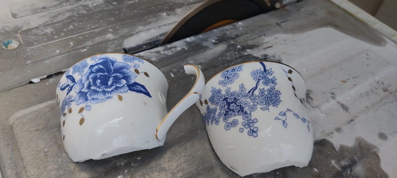 May include: Two broken white porcelain teacups with blue floral designs and gold trim. One cup features a large blue flower, while the other has a blue floral branch pattern. A circular saw blade is visible in the background.