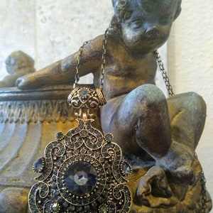 May include: A gold-tone necklace with a large, ornate pendant featuring a dark gray crystal surrounded by smaller crystals. The pendant is in the shape of a stylized floral design.