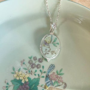 May include: A silver chain necklace with an oval pendant featuring a delicate floral design and a small bird. The pendant is made of white porcelain with a green and white floral pattern.