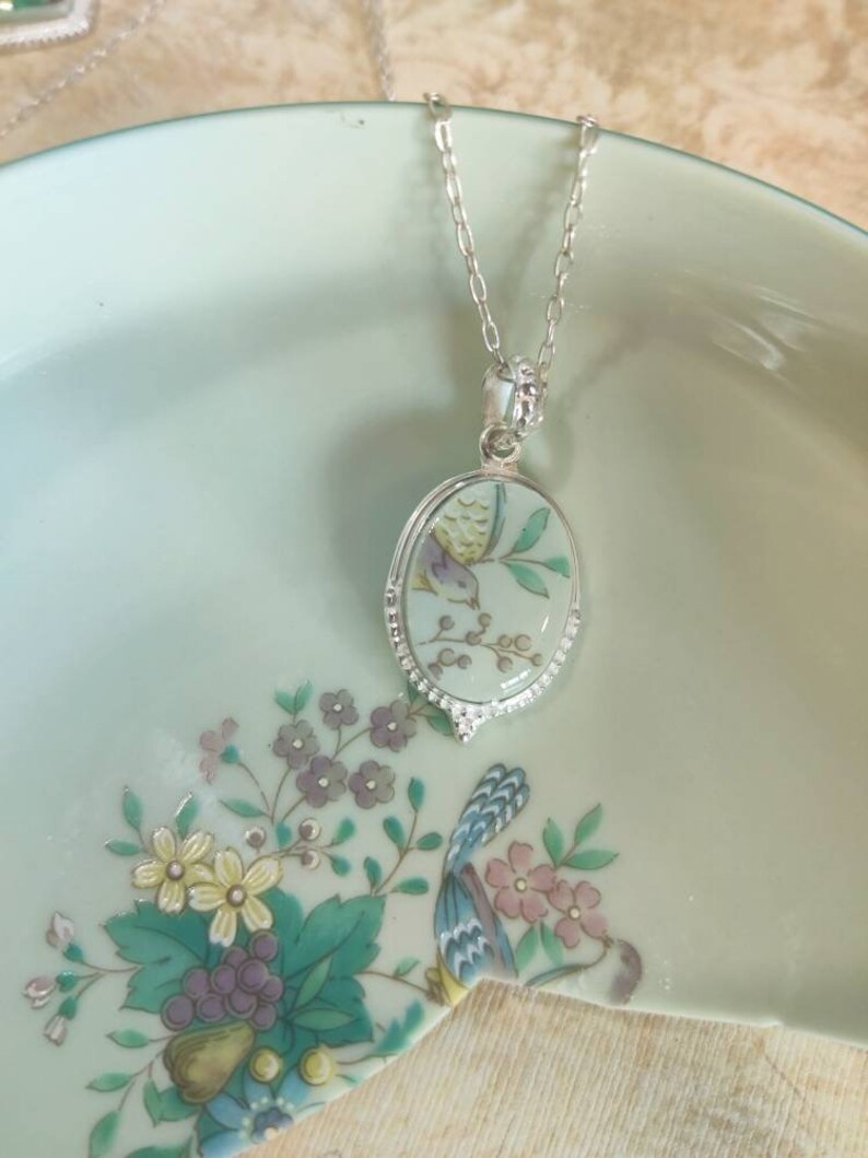 May include: A silver chain necklace with an oval pendant featuring a delicate floral design and a small bird. The pendant is made of white porcelain with a green and white floral pattern.
