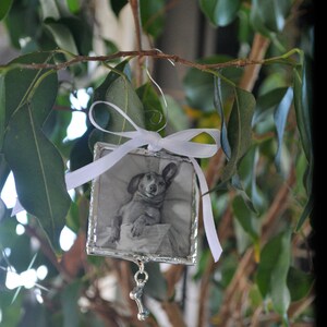 May include: A silver ornament with a white ribbon and a black and white photo of a dog inside. The ornament has a silver bone charm hanging from it.