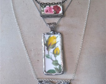Può includere: Quattro collane con ciondoli in vetro a motivi floreali. La collana superiore ha un ciondolo a forma di cuore con fiori rosa e gialli. La seconda collana ha un ciondolo rettangolare con fiori rossi e rosa. La terza collana ha un ciondolo rettangolare con fiori gialli. La collana inferiore ha un ciondolo a forma di cuore con fiori rosa e gialli.