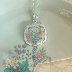 May include: A silver chain necklace with a square pendant featuring a hand-painted design of two birds and flowers on a light blue background.