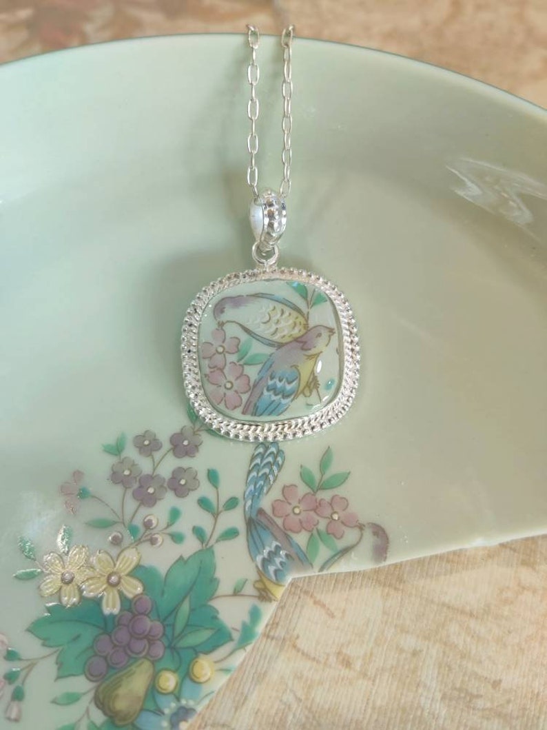 May include: A silver chain necklace with a square pendant featuring a hand-painted design of two birds and flowers on a light blue background.