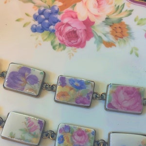 May include: A bracelet made of porcelain tiles with floral patterns. The tiles are connected by gold-toned metal links. The tiles feature a variety of flowers, including purple irises, pink roses, and yellow and blue wildflowers.