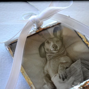 May include: A silver-framed ornament with a black and white photo of a dachshund dog wearing a sweater. The ornament is tied with a white ribbon.
