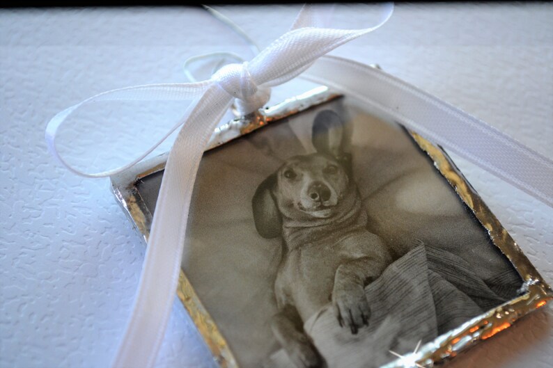 May include: A silver-framed ornament with a black and white photo of a dachshund dog wearing a sweater. The ornament is tied with a white ribbon.