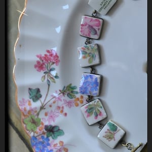 May include: A bracelet made with vintage porcelain tiles featuring floral patterns in shades of pink, blue, and green. The tiles are attached to a silver chain with a brass clasp. The bracelet has a tag that reads "Spode Copeland China England."